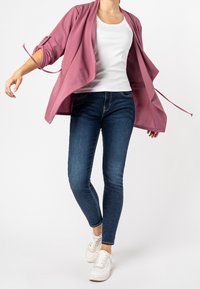 Veste ouverte rose poussiéreux, tissu léger, manches longues avec détail noué. Débardeur blanc en dessous, associé à un jean skinny bleu foncé et des baskets blanches.