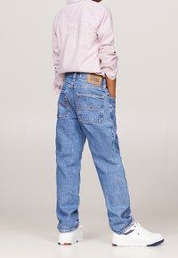 Persona vistiendo jeans claros de Tommy Hilfiger, camiseta de rayas de manga larga y zapatillas blancas, de pie con las manos en los bolsillos frente a un fondo liso.