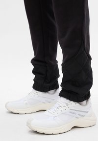 Białe siatkowe sneakersy z teksturowanymi akcentami i detalem logo, zestawione z czarnymi spodniami dresowymi z pikowanym dolnym fragmentem i elastycznymi mankietami.