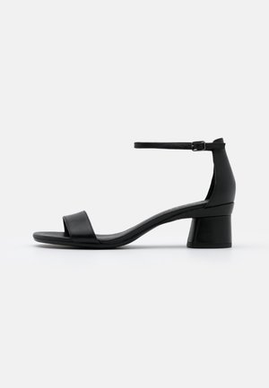 Sandale en cuir noir avec bout ouvert, bride à la cheville avec boucle et petit talon bloc sur fond blanc uni.