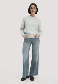 Hellgrüner, gestrickter Pullover mit Rundhalsausschnitt, kombiniert mit hellblauen, weit geschnittenen Jeans und dunkelbraunen Schuhen. Stehende Pose.