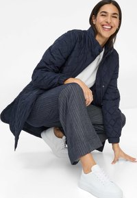 Veste matelassée marine avec un col haut, dotée de poignets élastiques et d'une fermeture à boutons. Associée à un pantalon gris à rayures et des baskets blanches.
