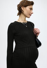 Μαύρο ριμπ φόρεμα με μακριά μανίκια και εφαρμοστή σιλουέτα, φορεμένο με στρωμένα χρυσά κολιέ και μια μικρή μαύρη τσάντα χειρός.