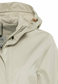 Veste beige clair avec capuche doublée en mesh, fermeture éclair, bouton-pression et cordon de serrage réglable au niveau du col.