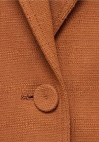 Tissu texturé brun mettant en valeur un revers et un bouton arrondi avec un détail de couture visible. Le matériau semble doux et tissé.