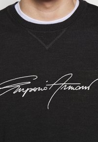 Black sweater featuring a textured knit, round neckline, and white embroidered signature accent on the front.