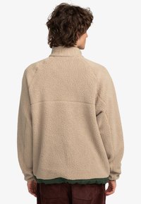 Beige fleece jacket with a high collar, textured surface, and raglan sleeves. Features a green hem and simple stitching detail.
