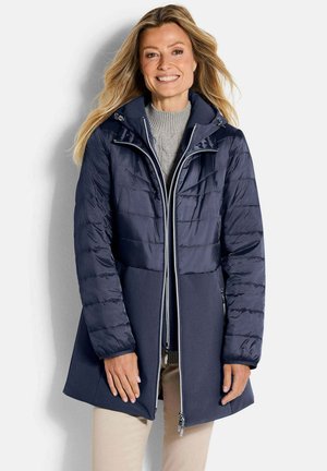 Frau lächelnd, die einen marineblauen gesteppte und wollgemischten Wintermantel über einem grauen Strickpullover und beigen Hosen trägt, steht vor einem schlichten Hintergrund.