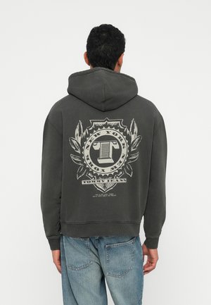 Muž v tmavosivej mikine s kapucňou s veľkým emblémom "Tommy Jeans New York" na chrbte, v kombinácii so svetlými modrými džínami, ktorý sa otáča chrbtom.