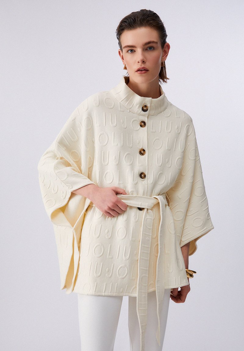 Poncho de textura cremosa con patrón en relieve del logo, grandes botones y cintura con cinturón. Presenta mangas anchas y cuello alto.