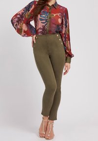 Vrouw die olijfgroene hoge taille skinny broeken met knoopdetails draagt, een meerkleurige blouse met lange mouwen en geplooide mouwen, en gouden strap-sandalen.