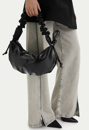 Kobieta trzymająca czarną torebkę z marszczonym uchwytem z napisem "JENNY", ubraną w jasnoszare, szerokie spodnie z bocznymi rozcięciami oraz czarne, spiczaste buty.