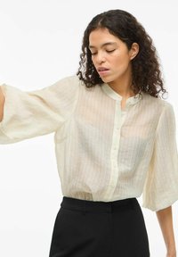 Blusa de crema ligera y transparente con rayas verticales, cierre de botones en la parte delantera y mangas sueltas y fluidas. Llevada con pantalones negros ajustados.