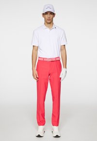 Witte polo, felroze broek, bijpassende riem en witte handschoenen. Casual schoenen met zwarte accenten. Schone, moderne atletische uitstraling.