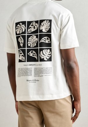 White t-shirt with black grid featuring nine stylized tropical leaves and text about nature and biodiversity on the back, worn by a person.