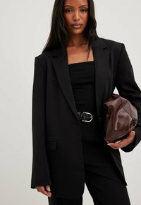 Must kandiline bleiser, millel on pikad varrukad, sälgud ja eestaskud; kombineeritud musta pluusi, musta vöö ja pruuni tekstuuriga kotti.