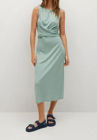 Robe midi sans manches vert clair avec un design à l'avant torsadé. Tissu doux et coupe décontractée, associée à des sandales bleu foncé.