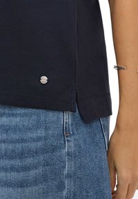Mørkeblå polo shirt i tekstureret stof, sølvlogo detalje ved kanten og sideslidser. Sættes sammen med lyseblå denim.