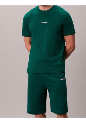 Homme portant un t-shirt à manches courtes Calvin Klein vert foncé et un short assorti, debout les mains derrière le dos sur un fond uni.