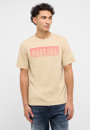 Junger Mann mit lockigem Haar, der ein beigefarbenes T-Shirt mit kurzen Ärmeln und rotem "MUSTANG"-Schriftzug sowie blaue Jeans trägt, vor weißem Hintergrund steht.