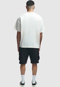 T-shirt blanche surdimensionnée à manches courtes, portée avec un short cargo noir et des baskets, vue de dos sur un fond neutre.