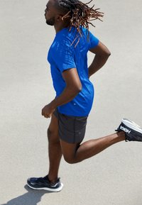 Blaues kurzärmliges Trainingsshirt und graue Shorts, die auf einer Oberfläche laufen, mit schwarzen Laufschuhen mit weißen Sohlen.