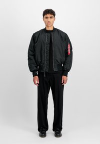 Jeune homme aux cheveux courts et bouclés portant une veste bomber noire tachetée, un pantalon noir et des chaussures noires, se tenant devant un fond clair uni.