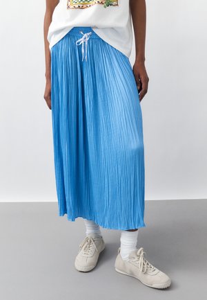 Άτομο που φοράει λευκό T-shirt με γραφικά, πλισέ midi φούστα σε έντονο μπλε με κορδόνι, λευκές κάλτσες και εκρού αθλητικά παπούτσια, όρθιο σε γκρι πάτωμα.