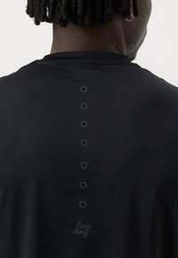 T-shirt de sport noir avec une texture lisse, sept petits boutons circulaires dans le dos et un logo discret dans le bas du dos.
