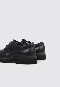 Chaussures en cuir noir à lacets avec semelle épaisse et col rembourré, inclinées pour montrer les détails arrière et latéraux sur un fond uni.