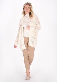 Cardigan a maglia traforata color crema, caratterizzato da una vestibilità ampia e rilassata con un motivo testurizzato; abbinato a pantaloni beige e scarpe di colore chiaro.