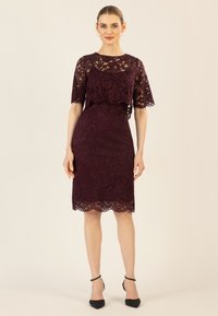 Spitzenkleid in tiefem Burgunderrot mit kurzen Ärmeln, gewelltem Saum und Figur betonter Silhouette. Schwarze High Heels ergänzen das Outfit.