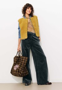 Un gilet en polaire jaune sur un haut à motifs, associé à un pantalon en velours côtelé large et foncé. Tenant un sac fourre-tout à carreaux marron avec un motif graphique.