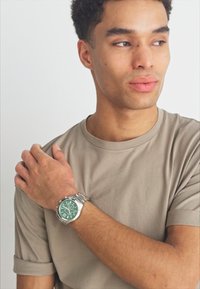 Silver watch with green dial and silver accents worn on dark beige t-shirt, showcasing round face, metal band, and clear hour markers.