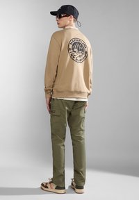 Felpa beige con logo nero sulla schiena, pantaloni cargo verdi e sneakers beige con dettagli. Un cappellino nero e occhiali da sole completano il look.