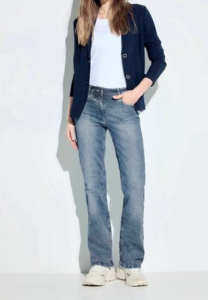 Frau trägt blauen Blazer, weißes T-Shirt, hellblaue Jeans und weiße Sneaker, steht mit einer Hand in der Tasche vor weißem Hintergrund.