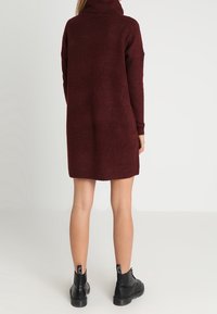 Vestito a maglione color bordeaux con colletto alto e maniche lunghe, che arriva a metà coscia. Abbinato a stivali neri con lacci alla caviglia.