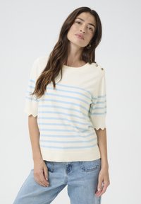 Pull crème avec des rayures bleu clair, bords des manches à volants et accents de boutons à l'épaule. Associé à un jean bleu clair.