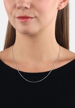 Donna che indossa una sottile collana d'argento sopra un top nero a coste, mostrando il collo, le clavicole e parte del viso inferiore.