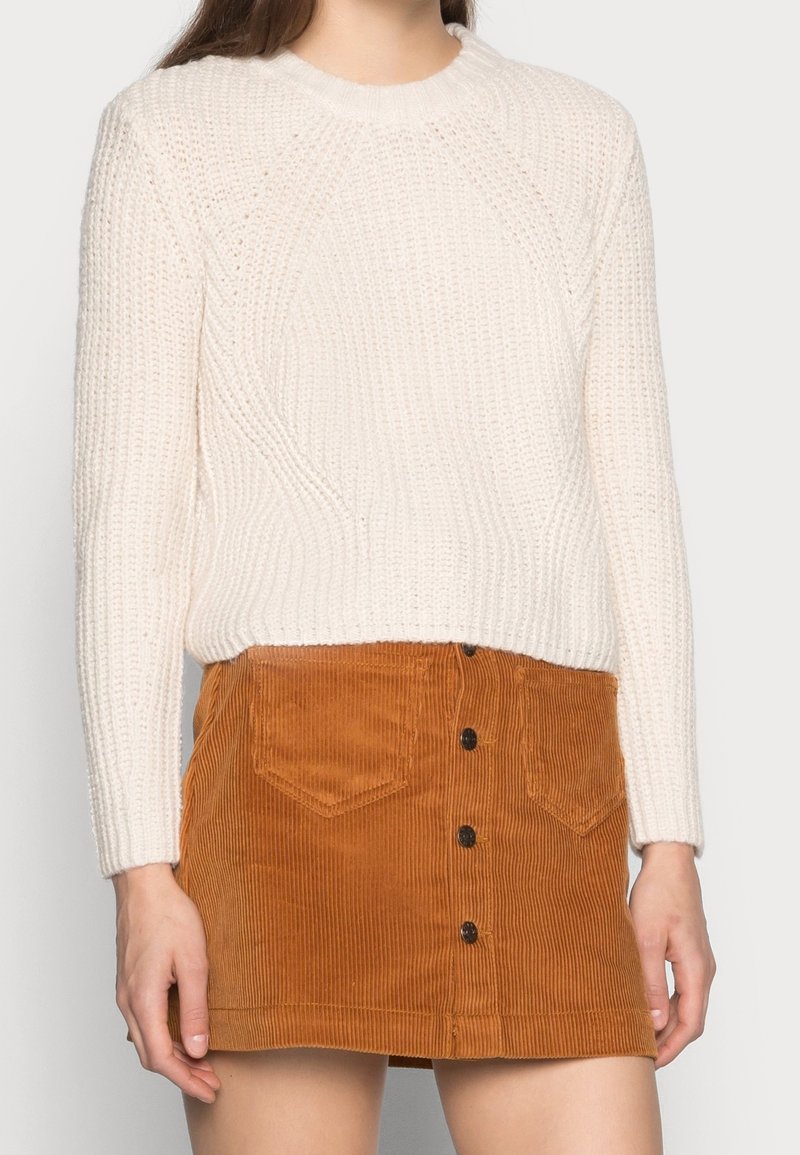 Femme portant un pull blanc à manches longues en maille et une jupe en velours côtelé marron avec des poches avant et des boutons sur le devant.