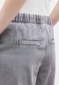 Κοντινό πλάνο ενός ατόμου που φοράει γκρι τζιν παντελόνι με ψηλή μέση, ελαστική μέση και τσέπες τύπου welt, σε συνδυασμό με λευκό τοπ.