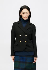 Dvoredno črn blazer z zlatimi gumbi in podrobnostjo na žepu, oblečen čez temno polo majico in karirano krilo v modrih in zelenih odtenkih.