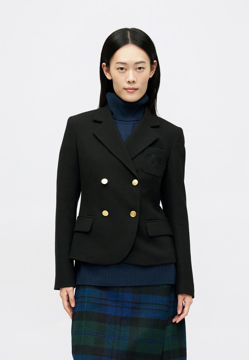 Dvoredno črn blazer z zlatimi gumbi in podrobnostjo na žepu, oblečen čez temno polo majico in karirano krilo v modrih in zelenih odtenkih.