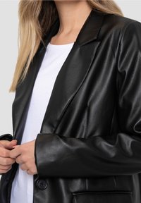 Blazer en simili cuir noir avec un col cranté et des manches longues, présentant une fermeture à un bouton et des poches latérales, porté sur une chemise blanche.