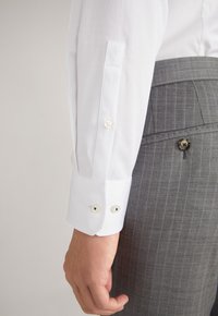 Puño de camisa blanca con dos botones, con un detalle bordado sutil. Pantalones grises con rayas finas parcialmente visibles en el encuadre.