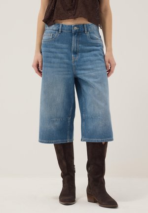 Pantaloncini di jeans blu a gamba larga di media lunghezza abbinati a stivali alti in suede marrone scuro e top corto marrone scuro.