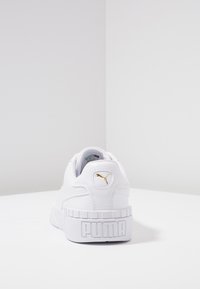 Zapatilla blanca con un acabado de cuero texturizado, presenta un logo dorado en el talón, una suela blanca gruesa y un acolchado alrededor del tobillo para mayor comodidad.