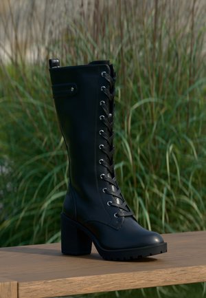 Botte noire à lacets montant jusqu’au genou avec un talon épais posée sur une surface en bois à l’extérieur, hautes herbes vertes floues en arrière-plan.