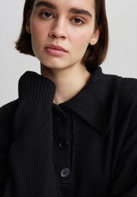 Cardigan nero a maglia con colletto e polsini a coste, dotato di quattro bottoni neri sul davanti. Texture liscia e vestibilità comoda.
