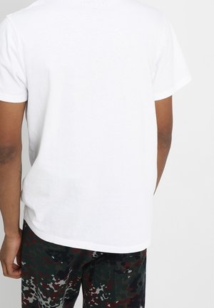 Person von hinten gezeigt, trägt ein schlichtes weißes T-Shirt mit kurzen Ärmeln und dunkle Tarnhosen vor einem hellgrauen Hintergrund.